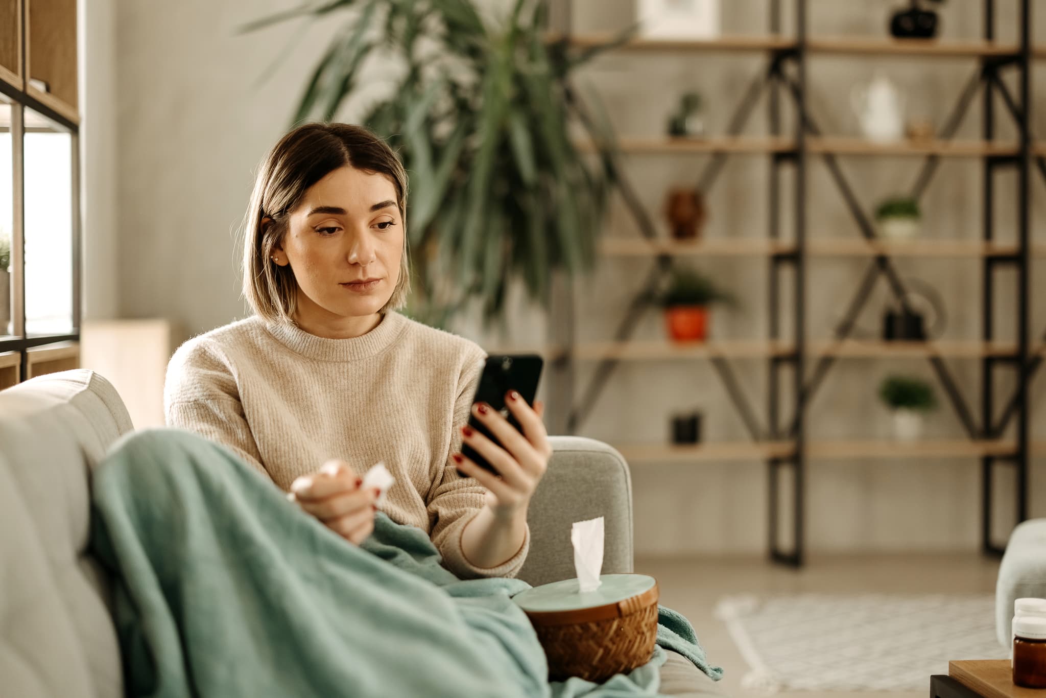 A woman sits on a couch wrapped in a green blanket, looking at her smartphone and holding a tissue, with a shelf and plants in the background.