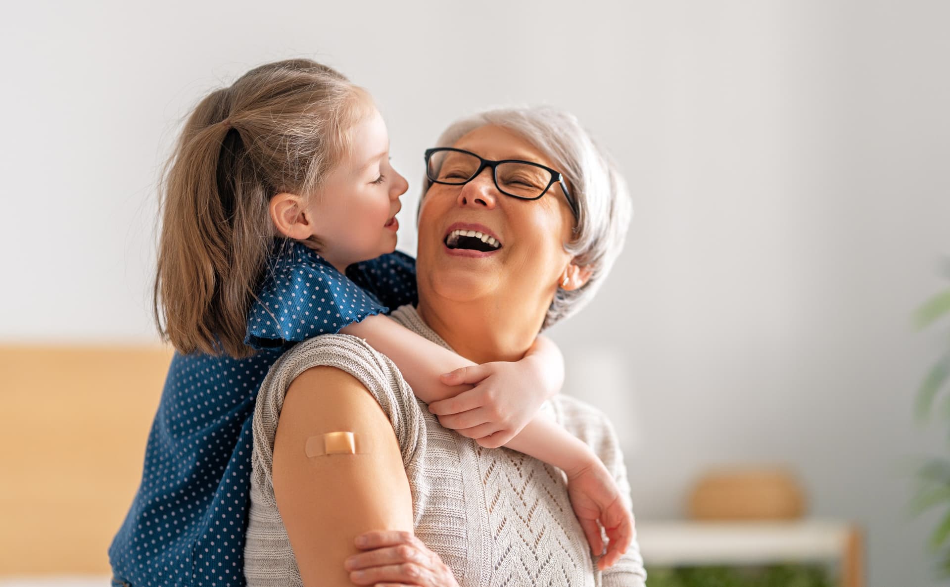 A joyful elderly woman with a bandage on her arm shares a happy moment with a little girl hugging her from behind.